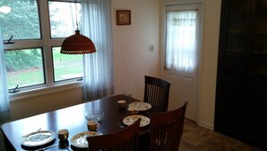 A dining area with a wooden table set for four, a hanging pendant light, and a window with sheer curtains. - Lake House Located Near North Fork Wineries (Southold)