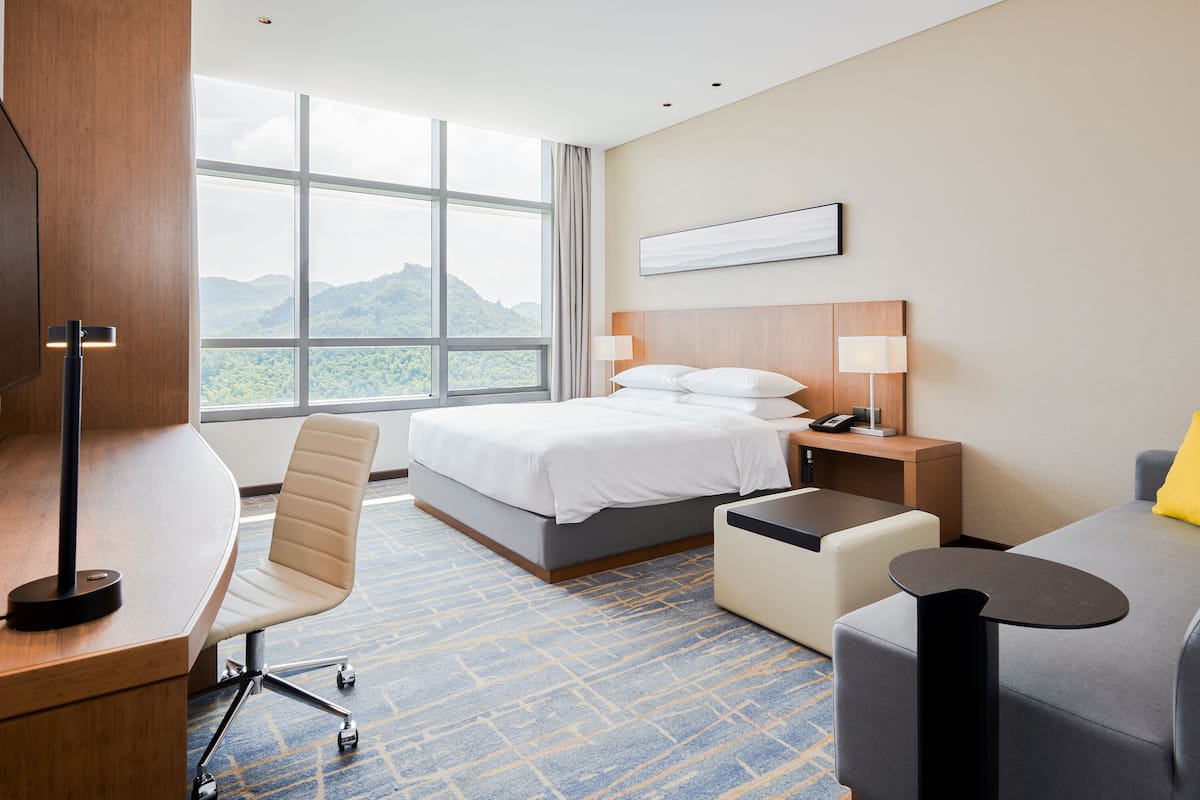 Una habitación de hotel con cama, escritorio, silla y vistas a montañas a través de ventanas amplias.