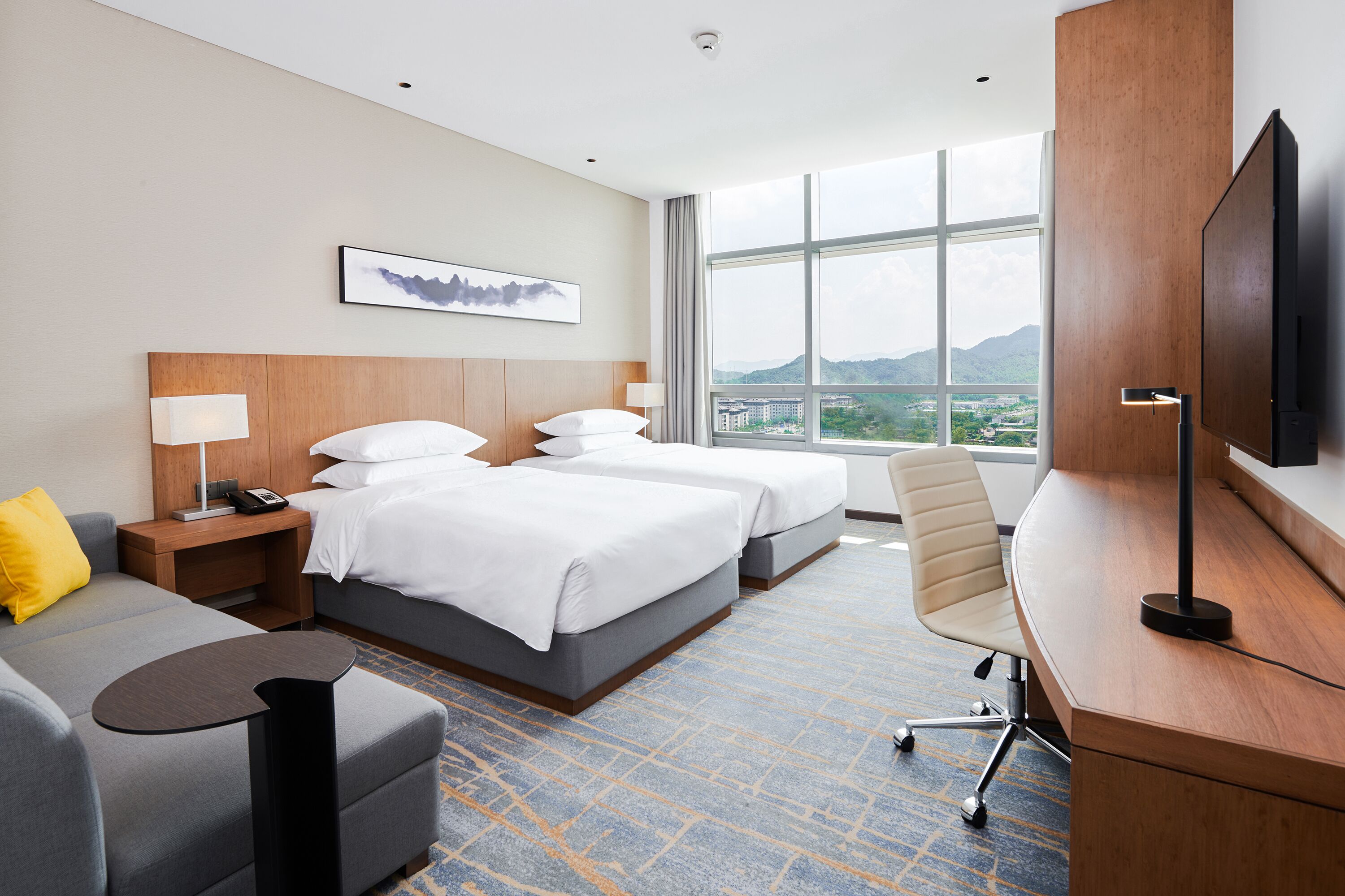 A hotel room with two beds, a desk, a chair, a sofa, a TV, and a large window offering a city view.