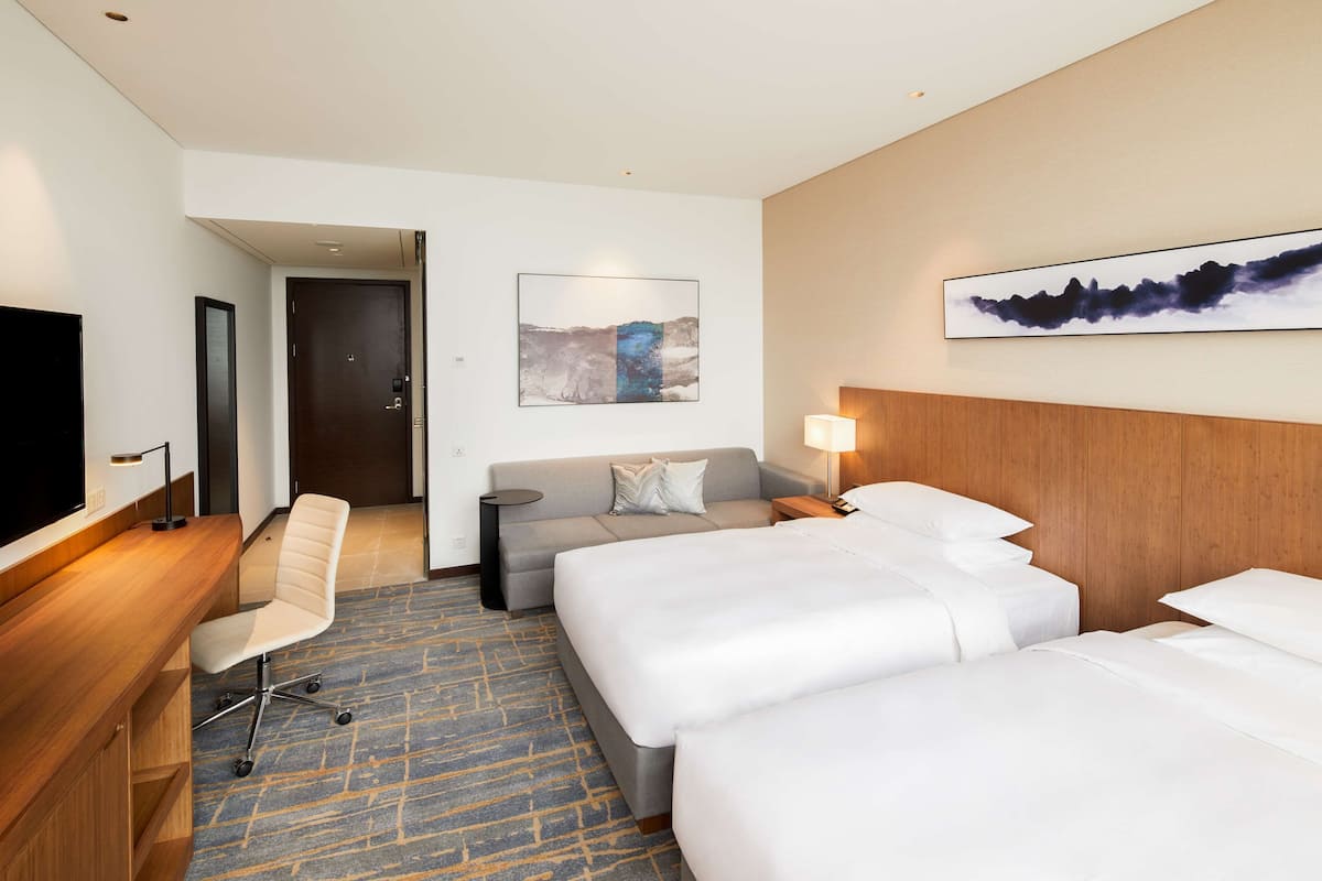Una habitación de hotel con escritorio, dos camas, sofá, televisión y obras de arte en las paredes.
