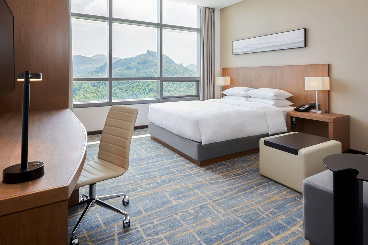 Habitación de hotel con cama, escritorio, silla, mesita de noche con lámpara y vistas a las montañas.