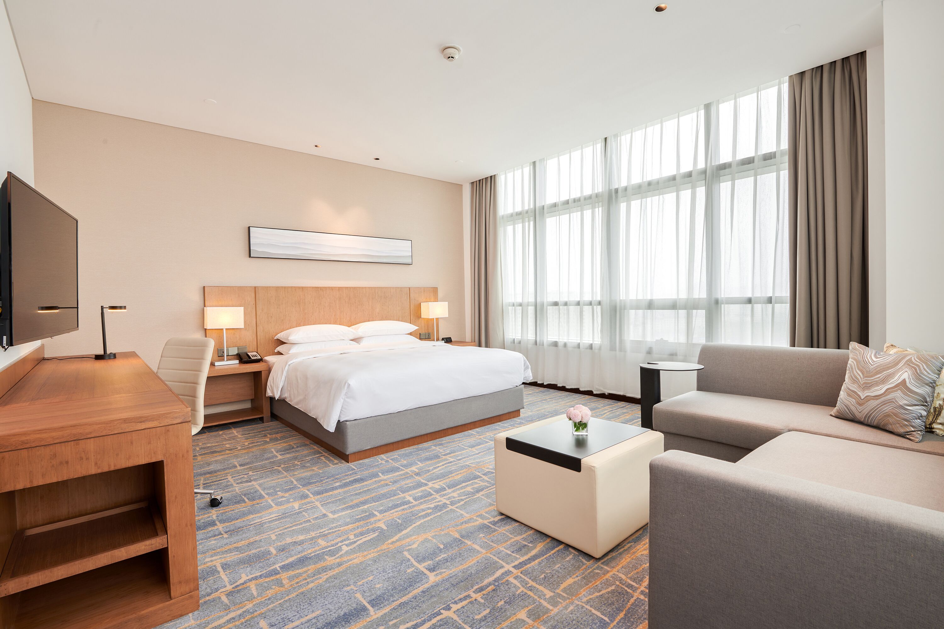 Una habitación de hotel con cama, sofá, escritorio, televisor y un ventanal con cortinas.
