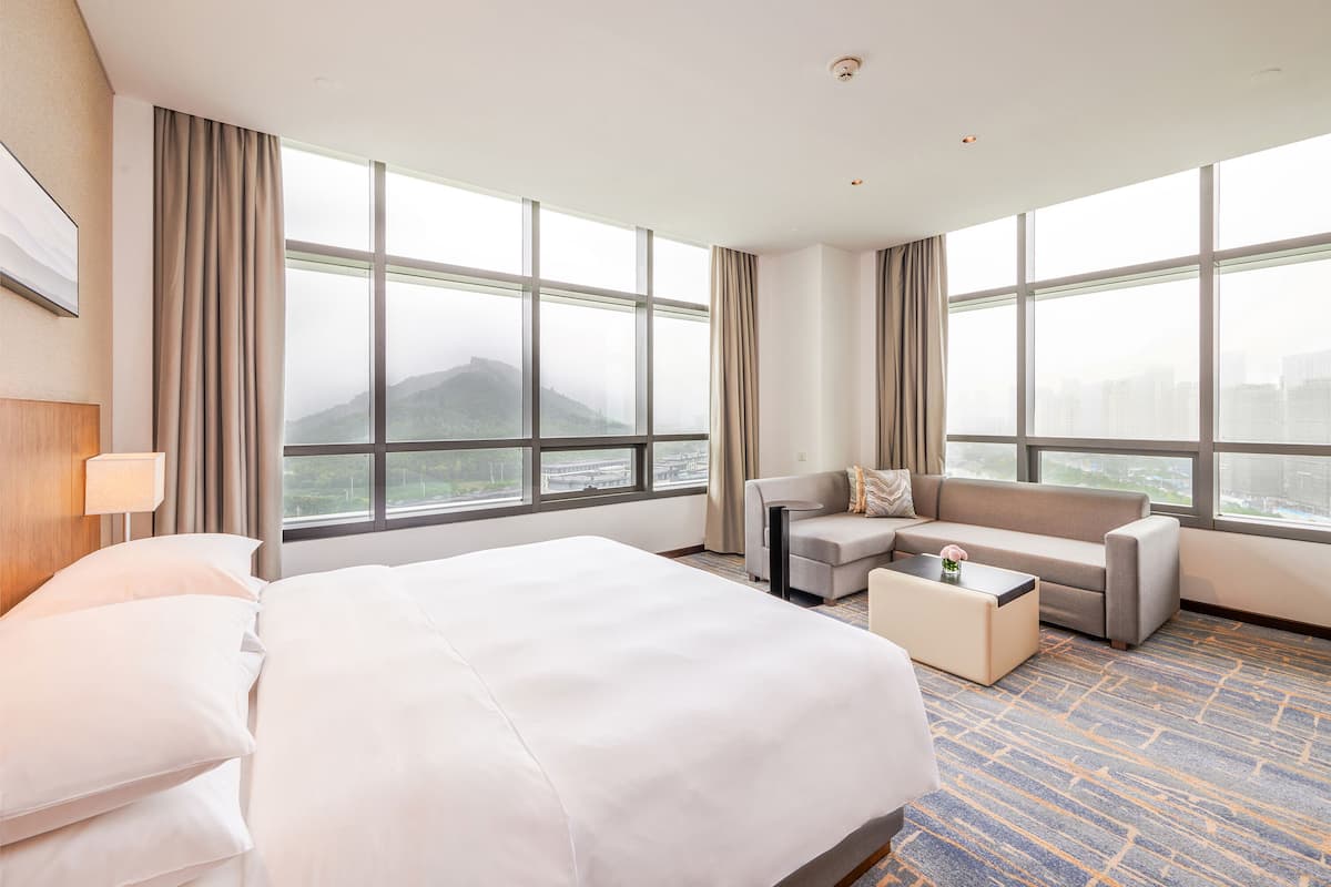 Habitación de hotel con una cama grande, un sofá, una mesa de centro y vistas a un paisaje urbano a través de amplias ventanas.