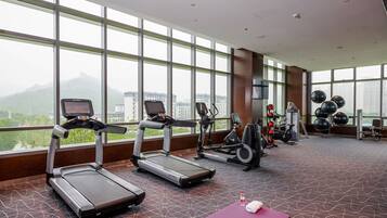 A gym with treadmills, stationary bikes, and weight machines, large windows with a city view, and a patterned floor.