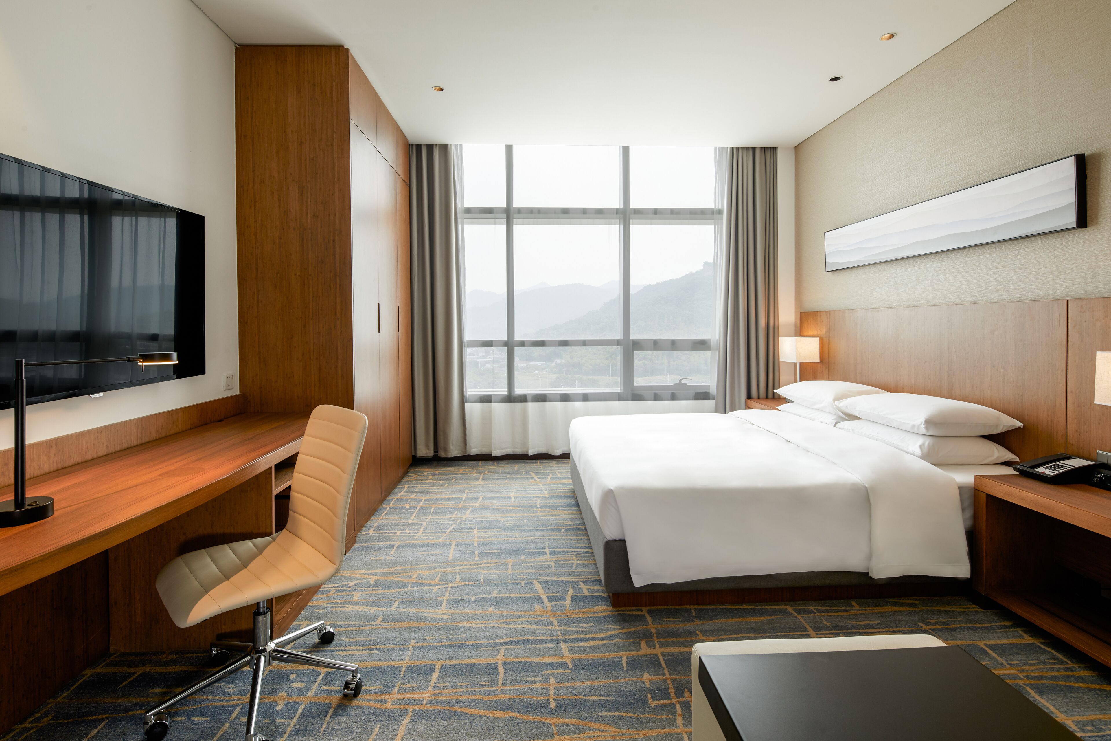 A hotel room with a bed, desk, chair, TV, and a view of the mountains through the window.