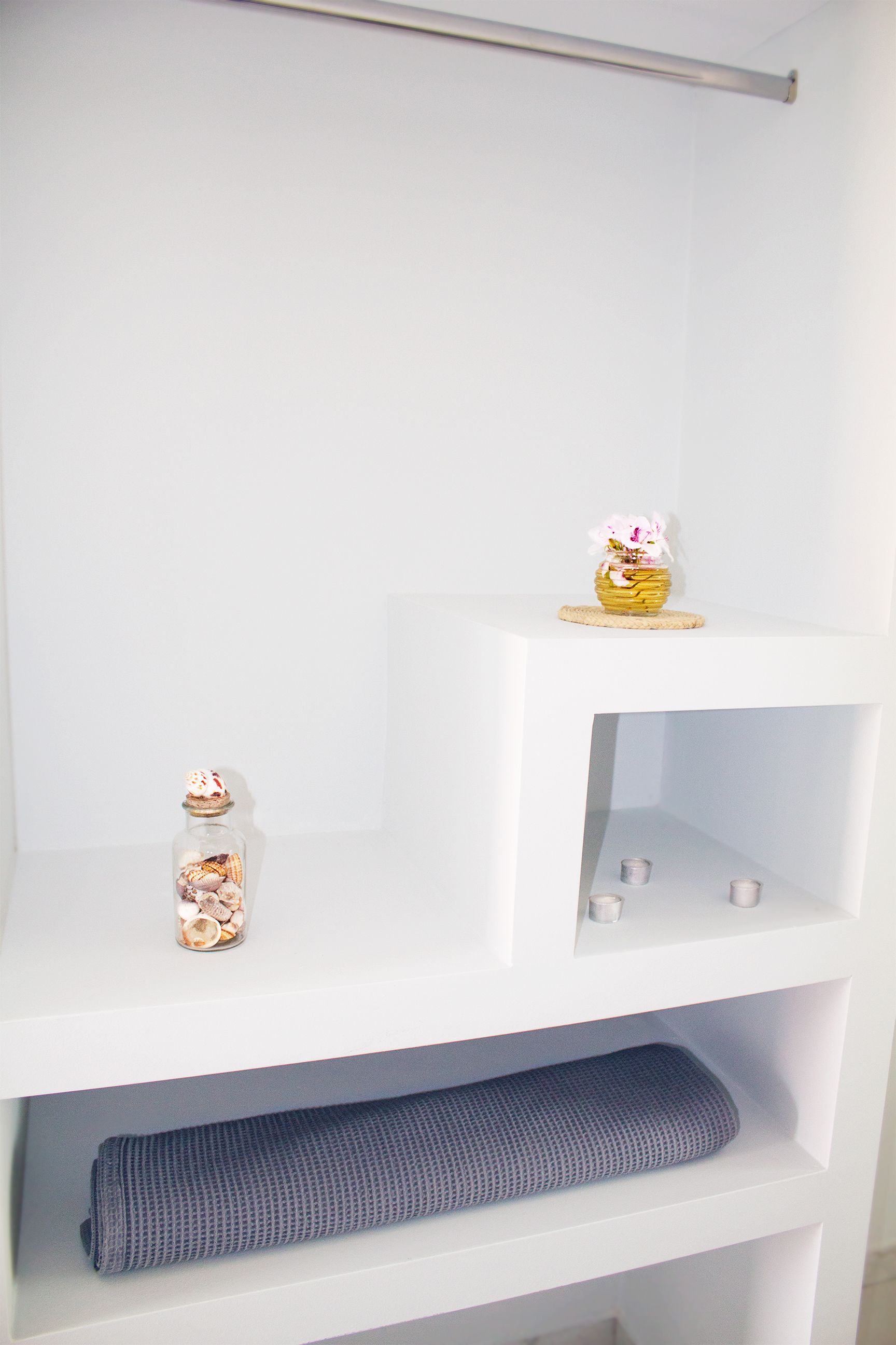 A white shelving unit with a basket of flowers on the top shelf, a glass jar with seashells on the middle shelf, and a rolled-up gray towel on the bottom shelf.