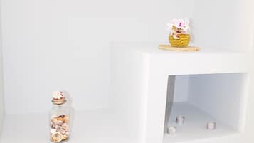 A white shelving unit with a basket of flowers on the top shelf, a glass jar with seashells on the middle shelf, and a rolled-up gray towel on the bottom shelf.
