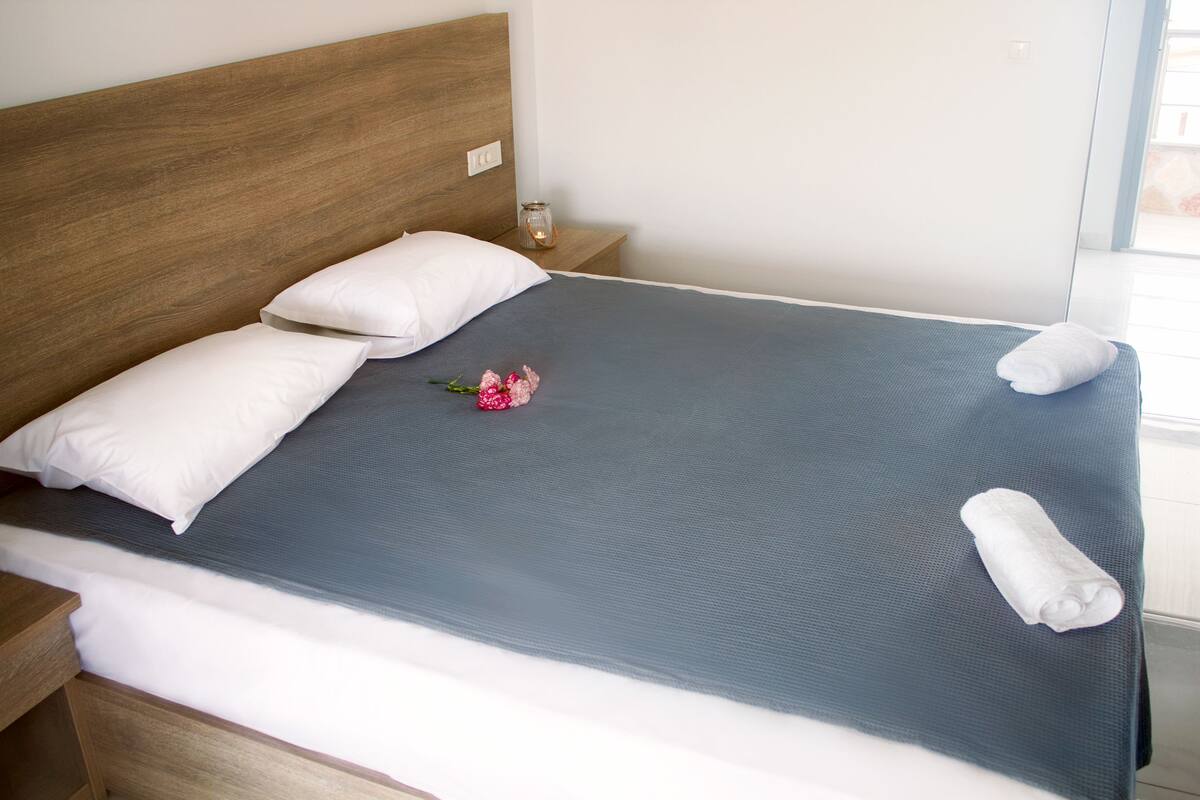 A neatly made bed with a blue bedspread, white pillows, and a rolled towel on the side.