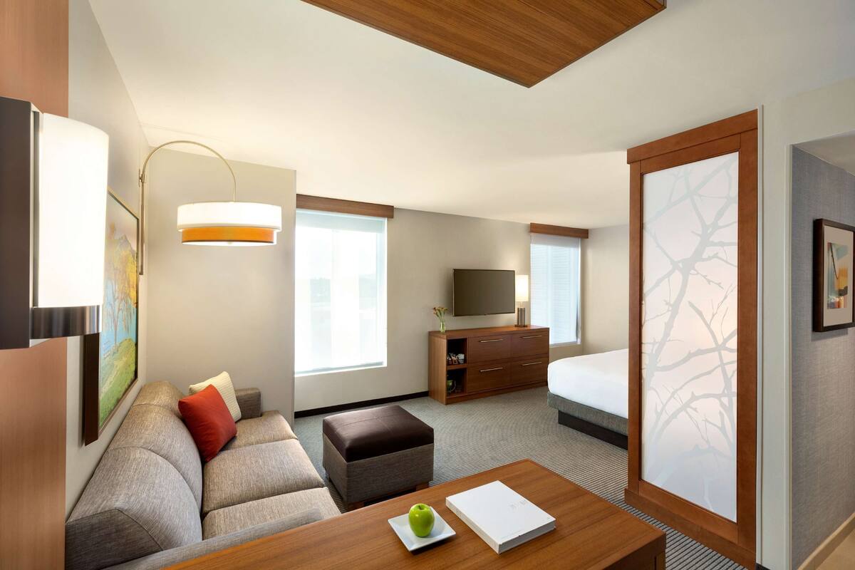 A modern hotel room with a sofa, ottoman, flat-screen TV, wooden dresser, and a large window.