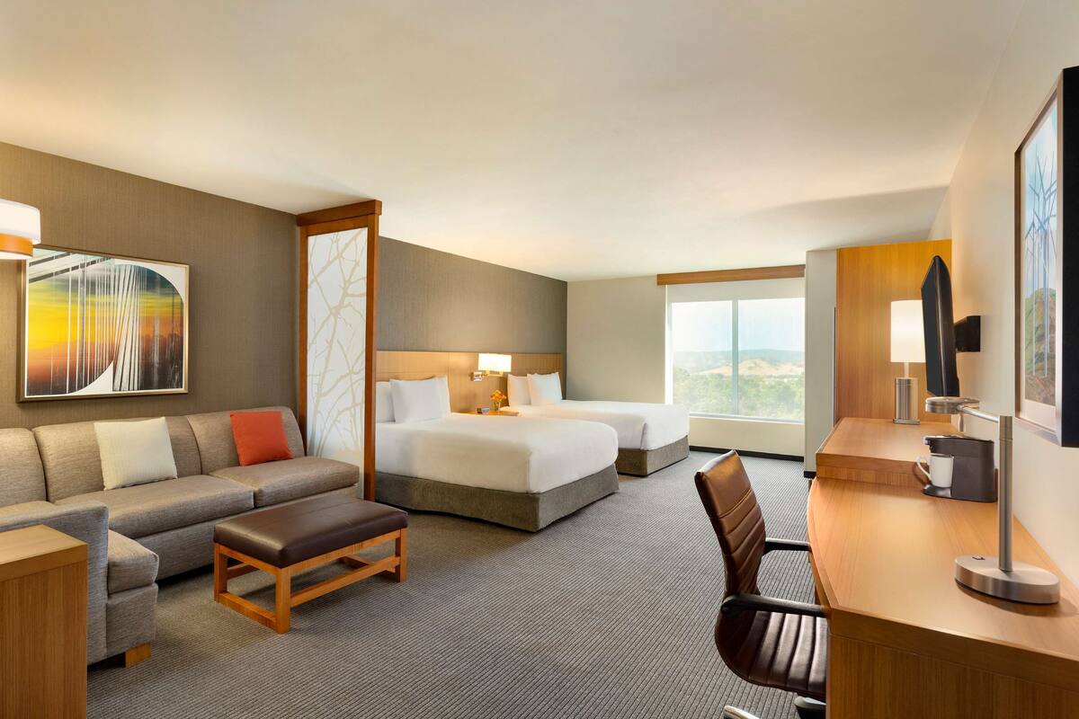 A hotel room with a sofa, two beds, a desk, and a large window offering a view.