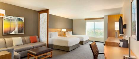 A hotel room with a sofa, two beds, a desk, and a large window offering a view.