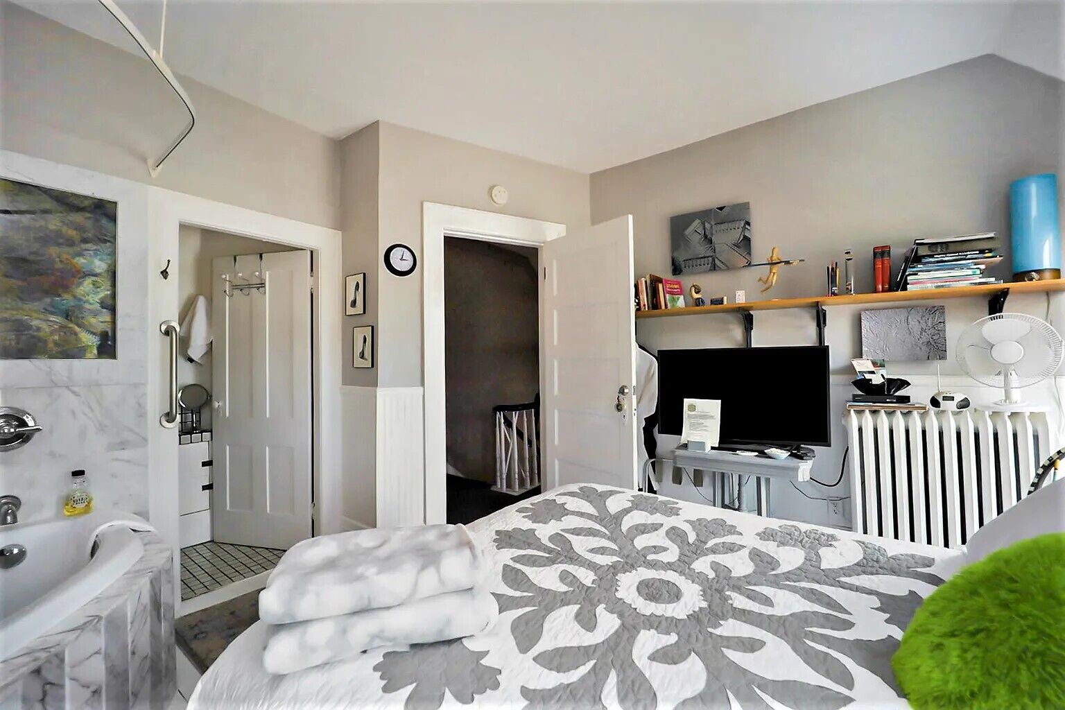A modern hotel room with a bathroom, a bed with a patterned bedspread, a television, a radiator, and a shelf with books and decorative items.