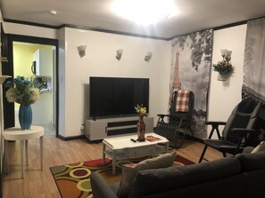 A modern living room with a flat-screen TV, a coffee table, a sofa, and a chair. There is a decorative vase on the coffee table and a painting on the wall. - Cozy French Pied a terre - King bed, washer/dryer (Schenectady)