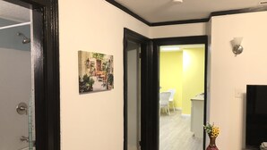 Un couloir avec un plancher en bois, un tableau représentant un jardin, une chaise blanche et un vase rempli de fleurs.
