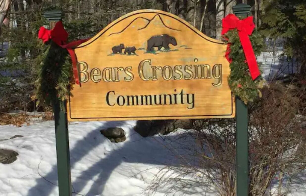 Un letrero de madera con un gráfico de un oso y montañas, que dice 'Bears Crossing Community', con lazos rojos en la parte superior.