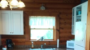 Een houten keukentje in een chalet, met een houten vloer, witte kastjes, een raam met gordijnen, een spoelbak, een fornuis en een koelkast.