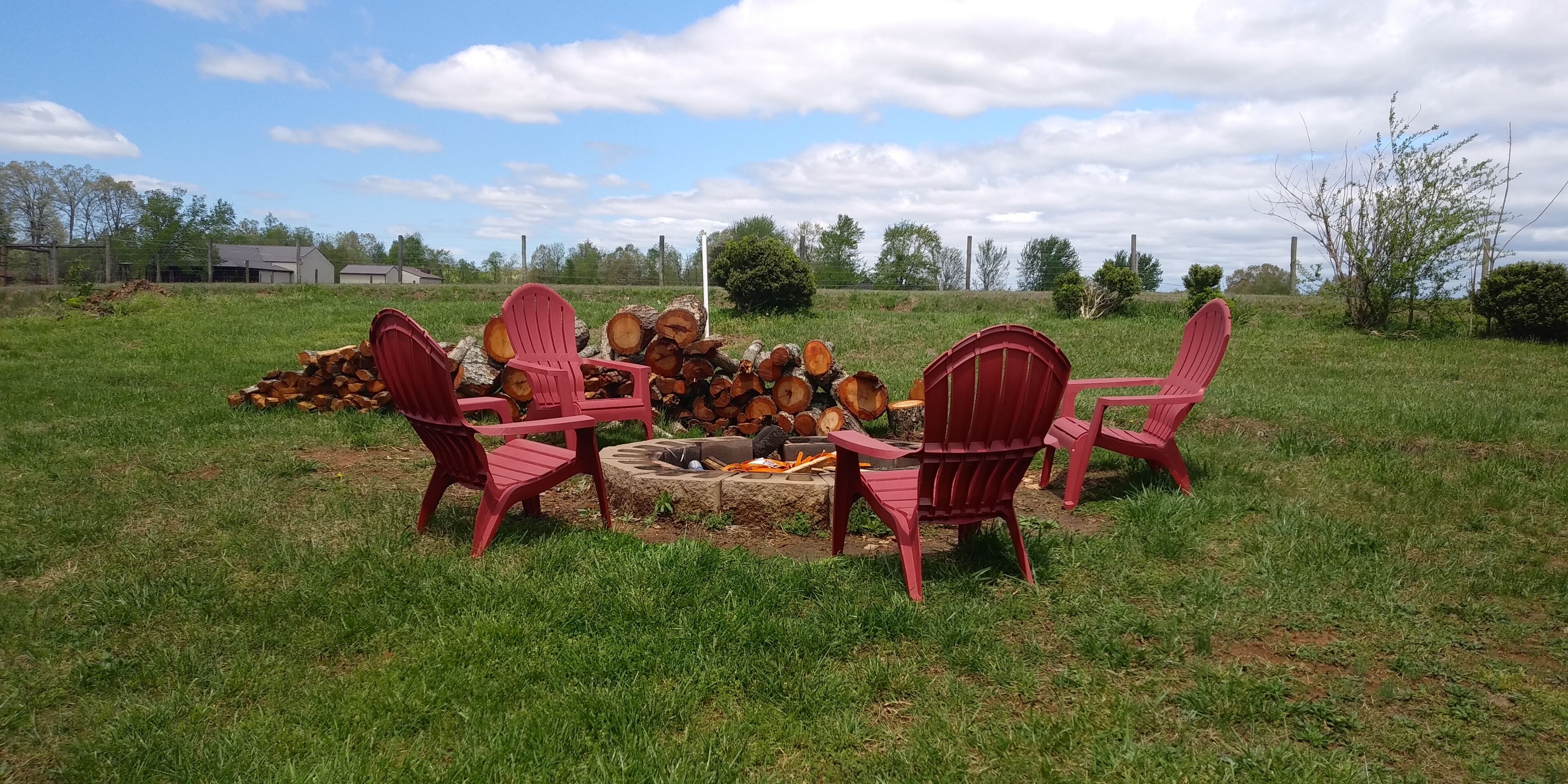 Eine Gruppe roter Adirondack-Stühle ist um eine Feuerstelle in einer grünen Außenanlage angeordnet.