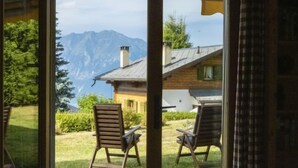 Een kamer met een groot raam dat uitzicht biedt op een berg en een houten huis. Er staan twee houten stoelen voor het raam.