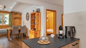 Een gezellige keuken met houten kastjes, een eethoek met stoelen, een koffieapparaat en uitzicht op een kamer met houten lambrisering.