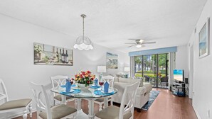 A dining area with a glass table, white chairs, and a ceiling fan. There is a painting on the wall and a view of the outdoors through large windows. - *Casa Mia* Fabulous 2BR Condo w/2Pools, Mins to IMG/Beach/Anna Maria Isl (Bradenton)
