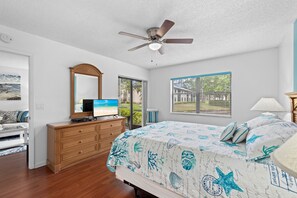 Ein Schlafzimmer mit Bett, Kommode, Deckenventilator, Fernseher und einem Fenster mit Jalousien.