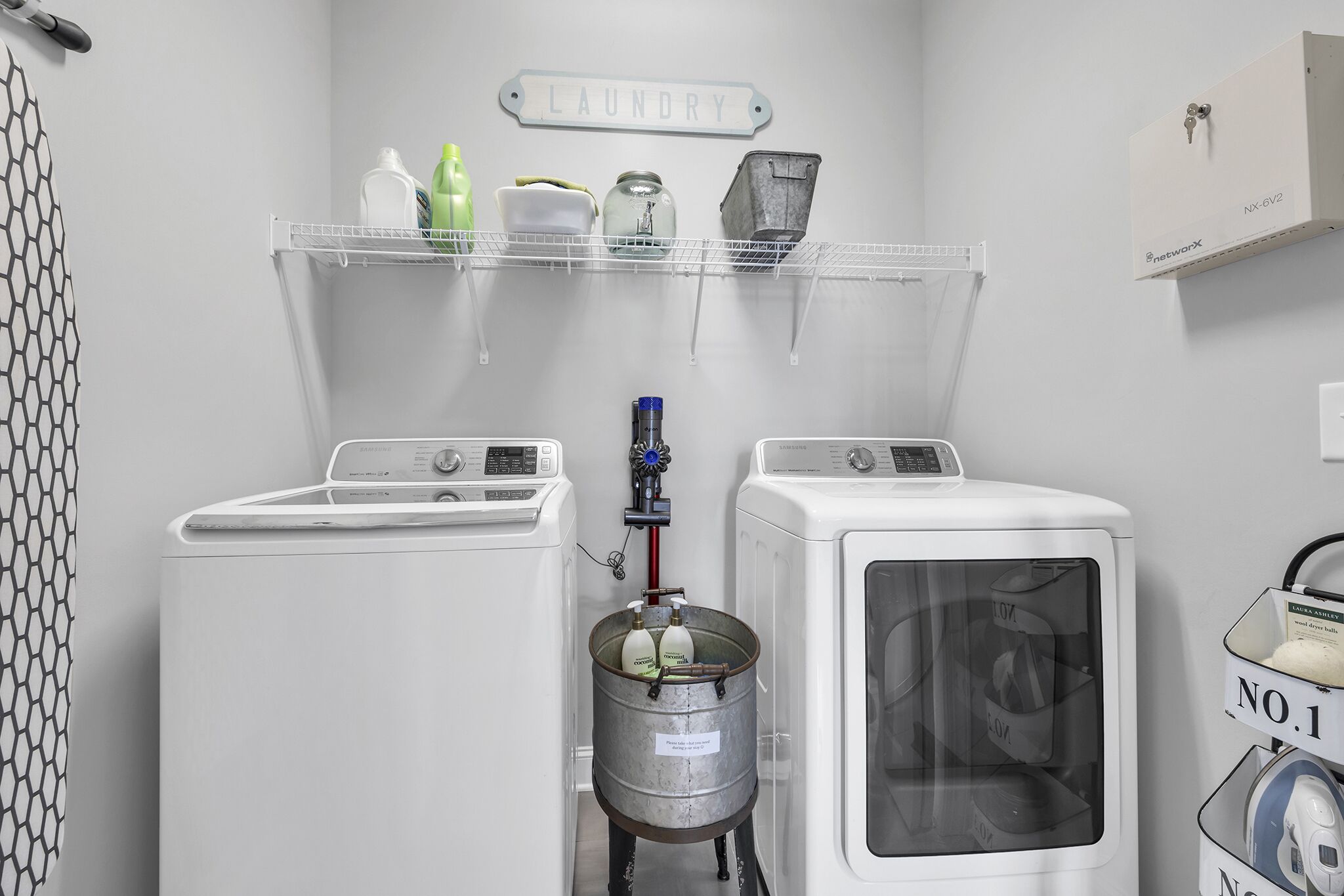 A laundry area with two washing machines, a metal bucket, and a shelf with cleaning supplies.