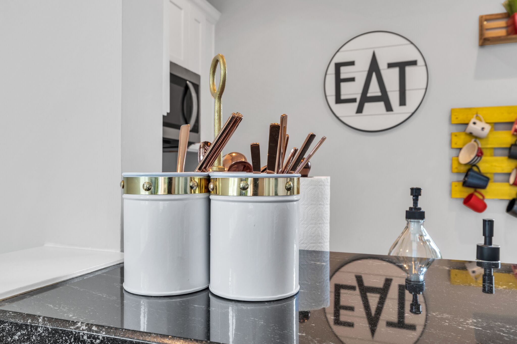 Eine Küchenarbeitsplatte mit zwei weißen Behältern, die Küchenutensilien, eine Glasflasche und ein an der Wand befestigtes Schild mit der Aufschrift 'EAT' enthalten.