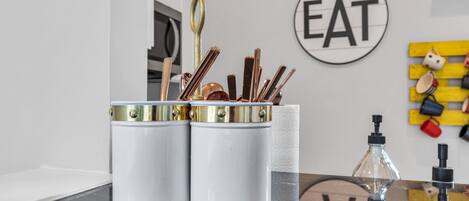 A kitchen counter with two white containers holding kitchen utensils, a glass bottle, and a wall-mounted sign that says "EAT".
