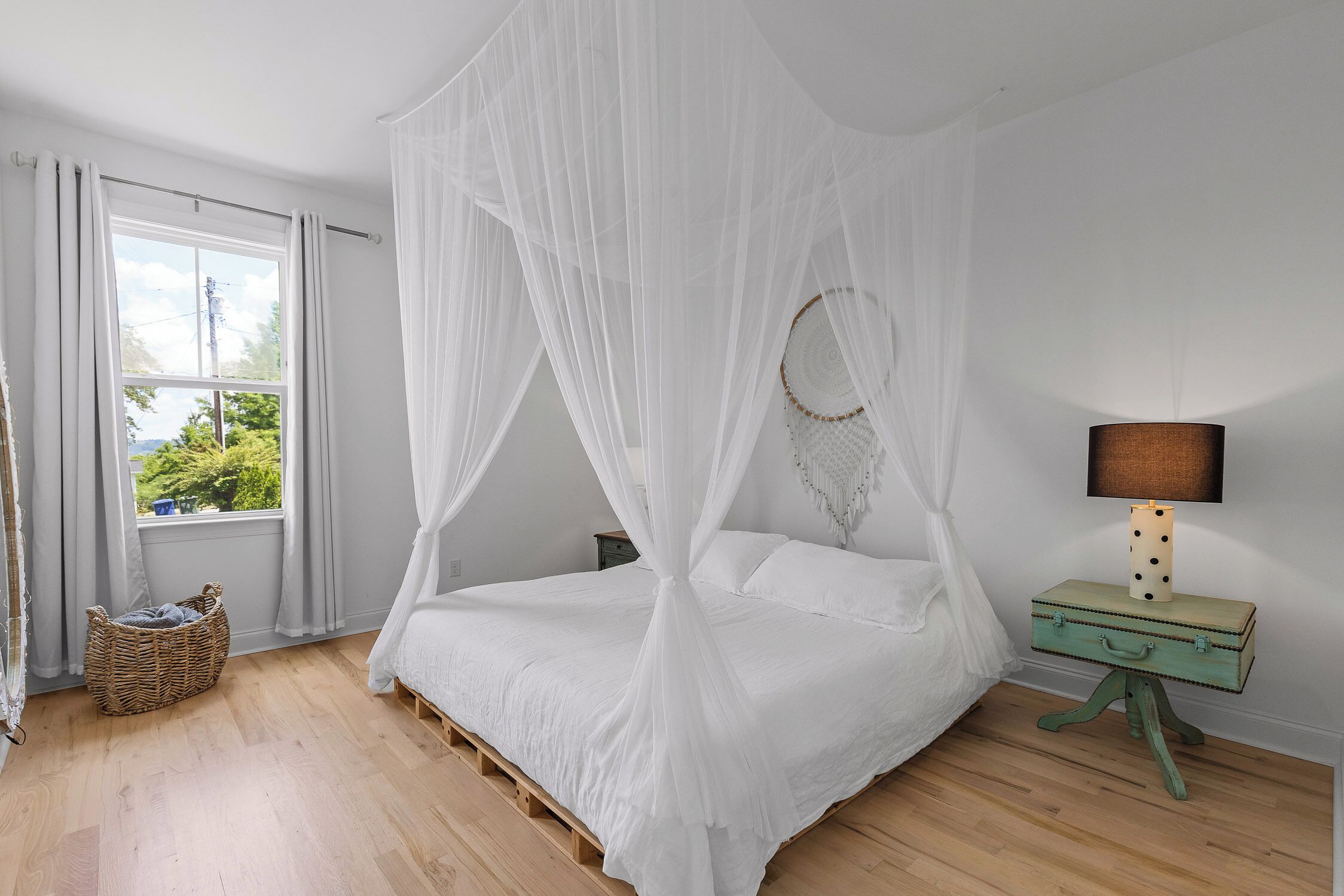 Un dormitorio con cama con dosel, una lámpara de noche, una cesta de mimbre y vistas al verde exterior.