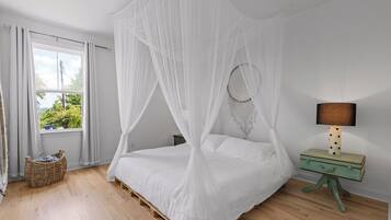 A bedroom with a four-poster bed, a bedside lamp, a wicker basket, and a view of greenery outside.