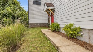 Fachada residencial con un camino de hormigón, un pequeño jardín y una casa con revestimiento blanco y puerta roja.