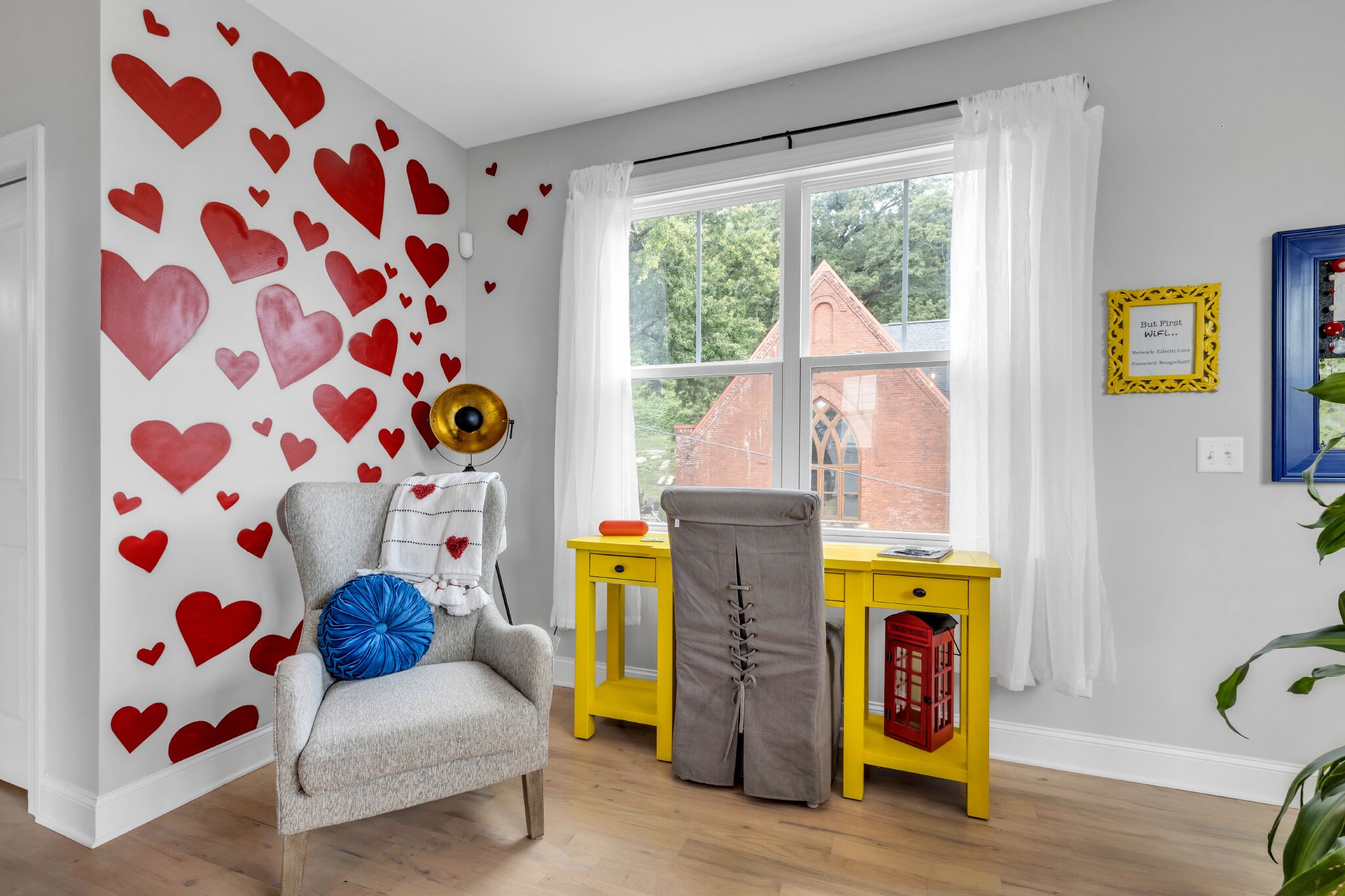 Una habitación con una pared blanca decorada con corazones rojos, un sillón gris con un cojín azul, un escritorio amarillo con un teléfono rojo y una ventana con cortinas blancas.