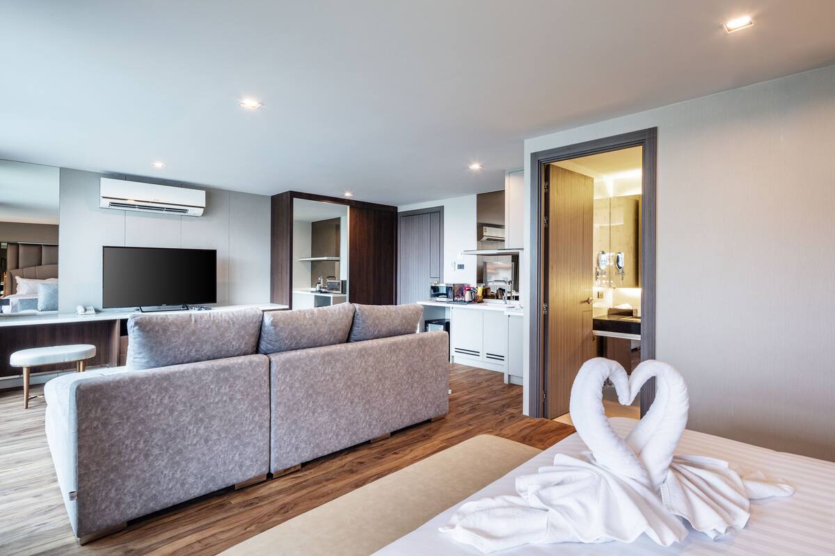 A modern hotel room with a sofa, a TV, a kitchen area, and a heart-shaped towel on the bed.