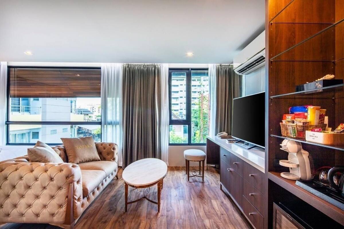 A modern hotel room with a sofa, a small round table, a TV, and a kitchenette area.