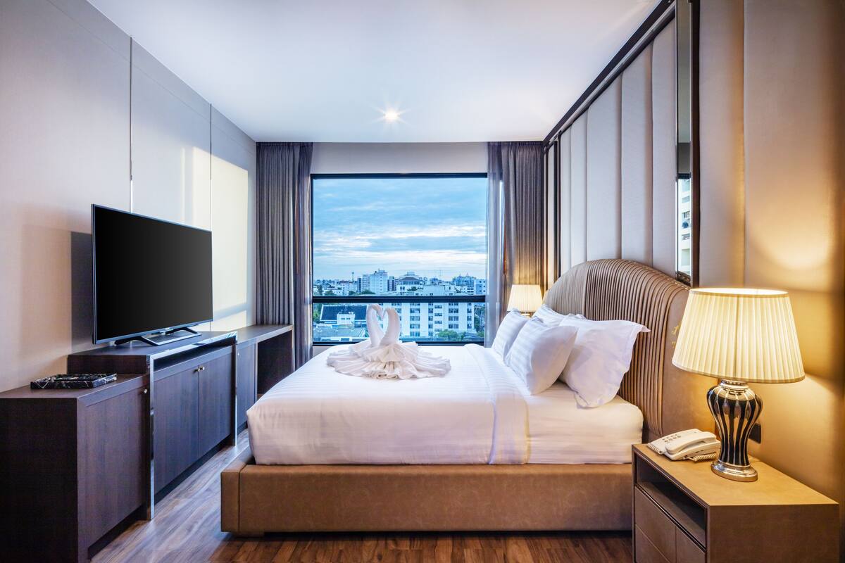 A hotel room with a large bed, a flat-screen TV, a bedside lamp, and a view of the cityscape.