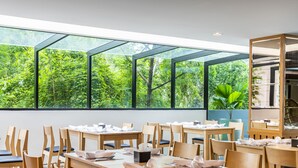 A restaurant with large windows offering a view of lush greenery. Tables are set with white tablecloths and napkins. - Grand Sala Thai (Bangkok)