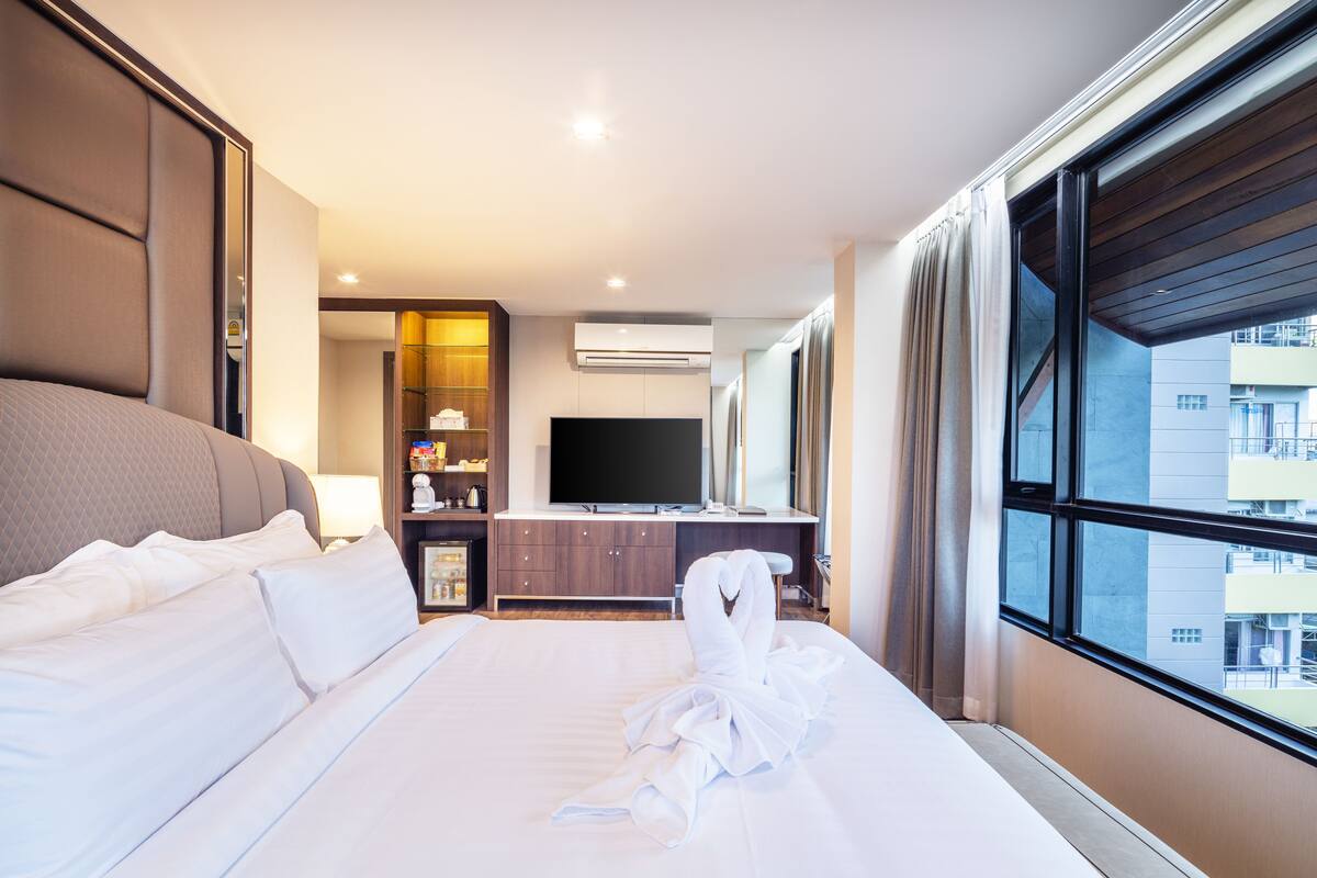A hotel room with a large bed, a TV, a desk, and a view of the city through the window.