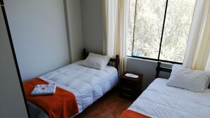 A hotel room with two single beds, each with white bedding and orange throw blankets. There is a small wooden nightstand with a lamp and a glass of water. The room has a window with curtains, and the floor is wooden. - Departamentos Arequipa Inn (Arequipa)