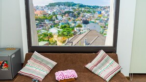 Una habitación con un gran ventanal que da a un paisaje urbano, un sofá marrón con cojines estampados y una pequeña mesa con refrigerador.
