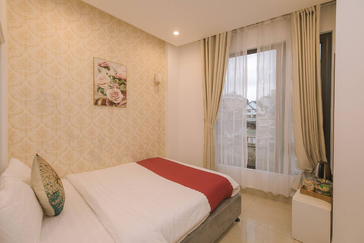 Een net opgemaakt bed met een rode deken, een bloemenpatroon op de muur en een raam met gordijnen.