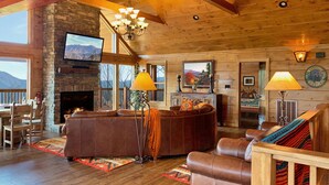 L’intérieur chaleureux d’un chalet en bois, avec une cheminée en pierre, des canapés en cuir, un coin repas et une grande fenêtre donnant sur un paysage montagneux.