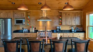 Une cuisine de style rustique, avec des armoires en bois, un réfrigérateur en acier inoxydable, un four à micro-ondes, une cuisinière et une machine à café. Trois lampes suspendues sont fixées au-dessus de l’îlot central, et une table avec quatre chaises se trouve à proximité.