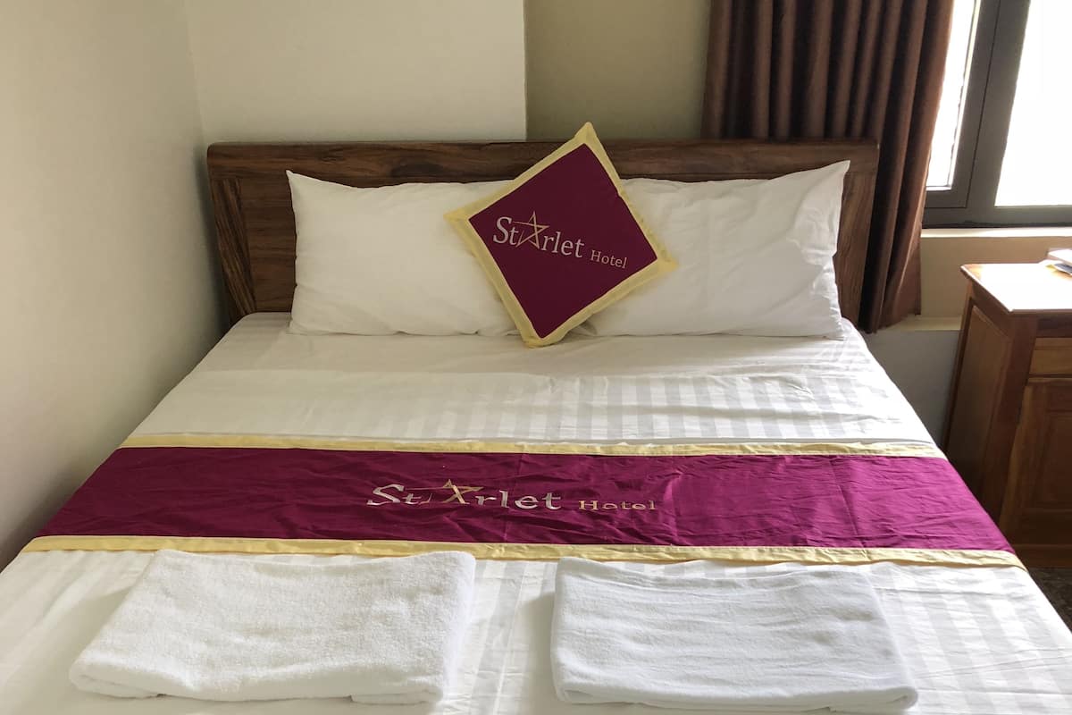 Una cama bien hecha con una colcha blanca y granate, dos toallas blancas dobladas y una almohada con el nombre del hotel.