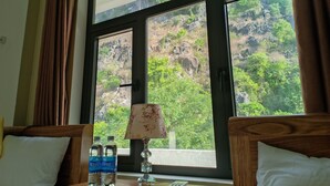 A hotel room with a large window offering a view of a mountainous landscape. There is a bed with two pillows and a bedside table with two water bottles and a lamp. - Starlet Hotel (Phong Nha)
