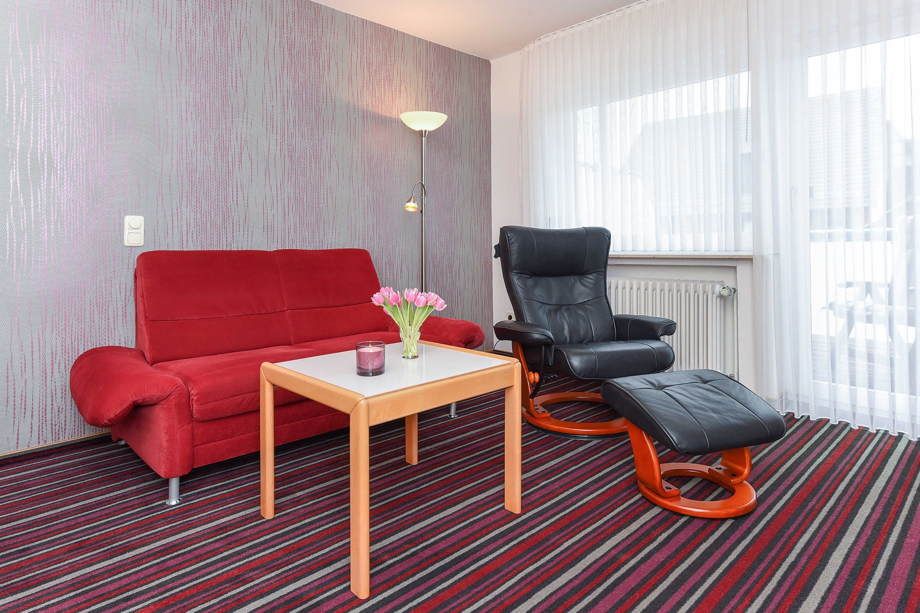 Una habitación con un sofá rojo, una silla negra, una mesita con un florero y una alfombra a rayas.