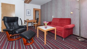 Moderni hotellihuone, jossa on musta nahkainen nojatuoli, punainen sohva, puinen sohvapöytä ja lattiavalaisin.