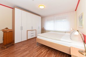 Une chambre avec un plancher en bois, un lit avec une literie blanche, une armoire, une table de chevet avec une lampe et une fenêtre avec des rideaux légers.
