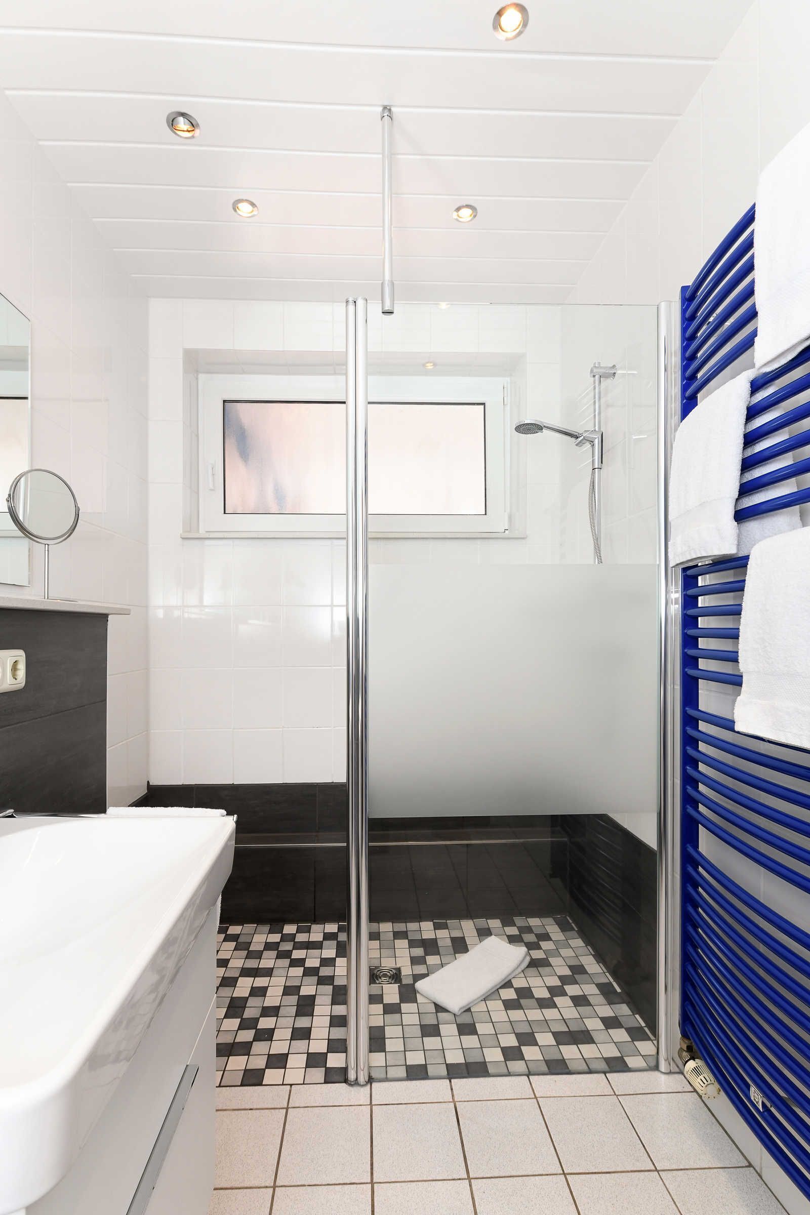 Un baño moderno con cabina de ducha de vidrio, una bañera blanca, piso a cuadros blanco y negro, y un perchero azul.