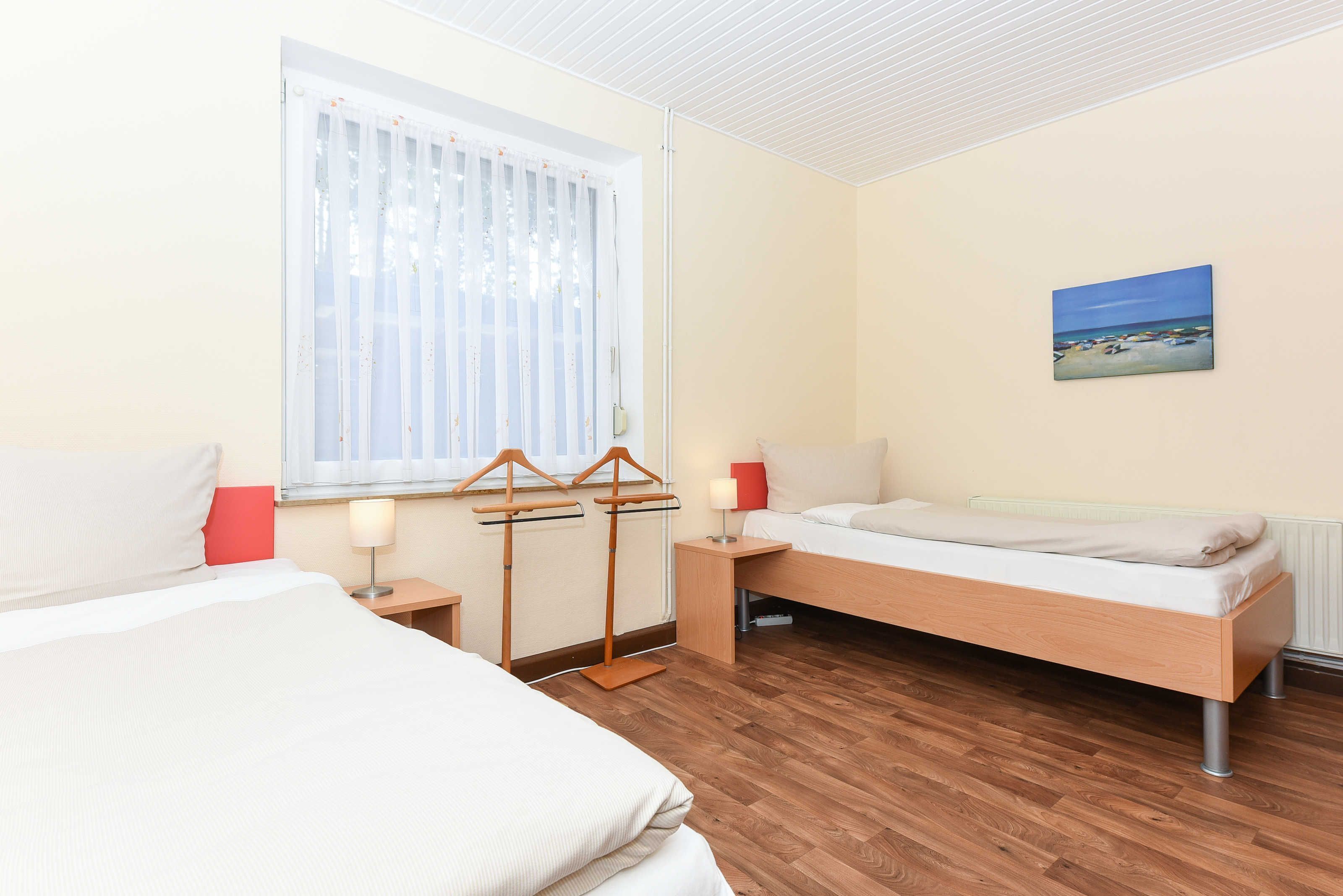 Habitació d'hotel amb dos llits, sòl de fusta, finestra amb cortines transparents, una tauleta de nit amb una làmpada i un quadre a la paret.