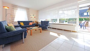 Een ruime woonkamer met een blauwe bank, een houten salontafel en een blauwe fauteuil. Er is een groot raam met dunne gordijnen, een radiator en uitzicht op het exterieur.